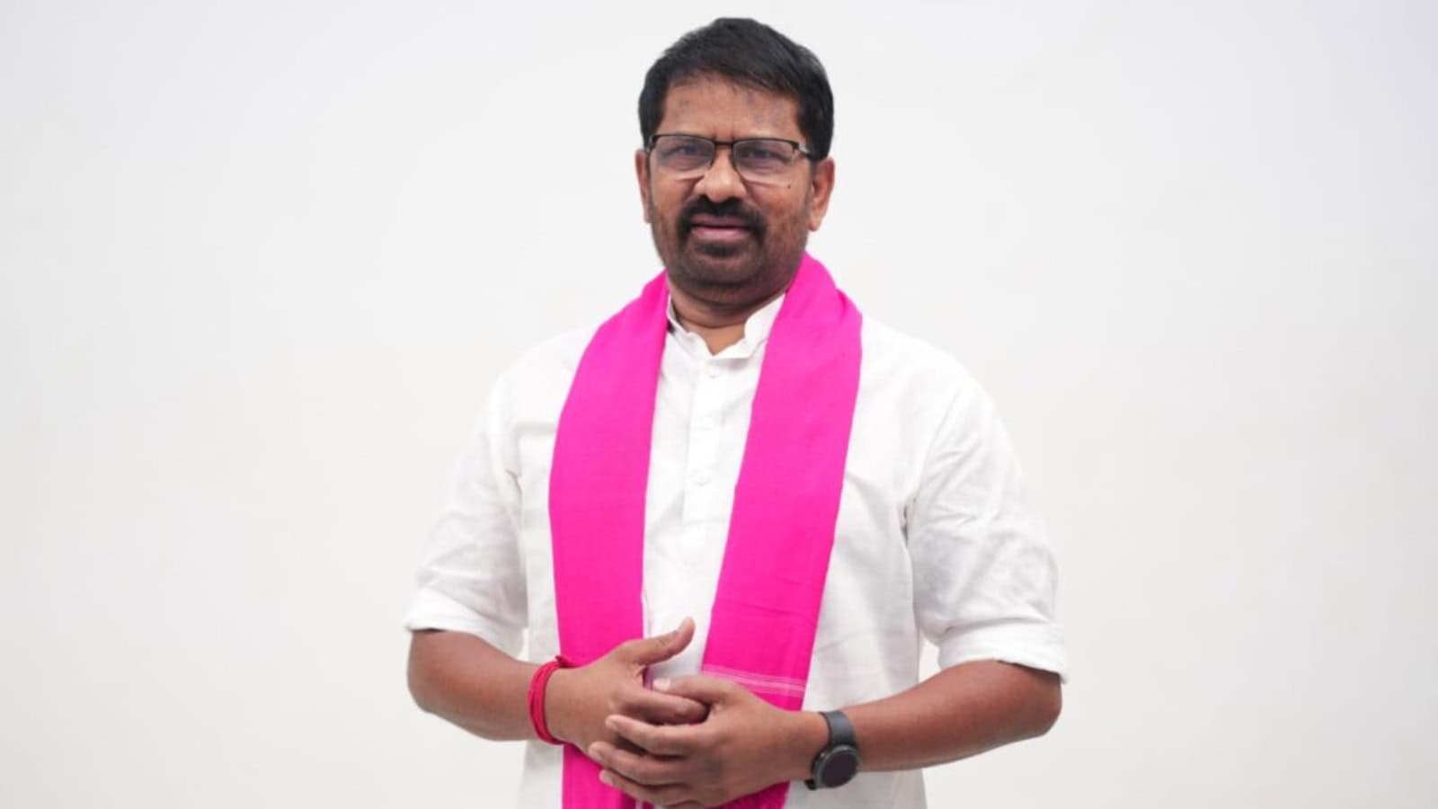 A man wearing glasses and a white shirt with a pink scarf, standing against a plain background.