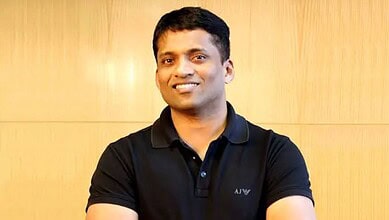 Portrait of a smiling Indian man in casual black polo shirt, standing against a wooden background.