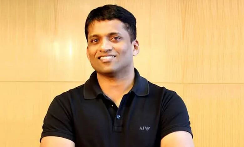 Portrait of a smiling Indian man in casual black polo shirt, standing against a wooden background.