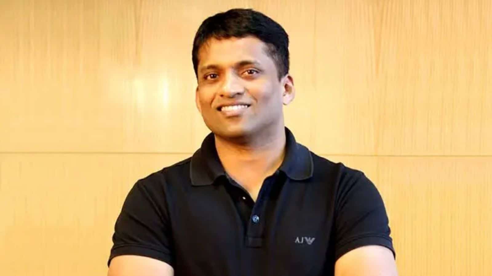 Portrait of a smiling Indian man in casual black polo shirt, standing against a wooden background.