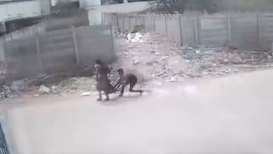 People walking along a dusty, unpaved road with scattered debris and fencing in the background.
