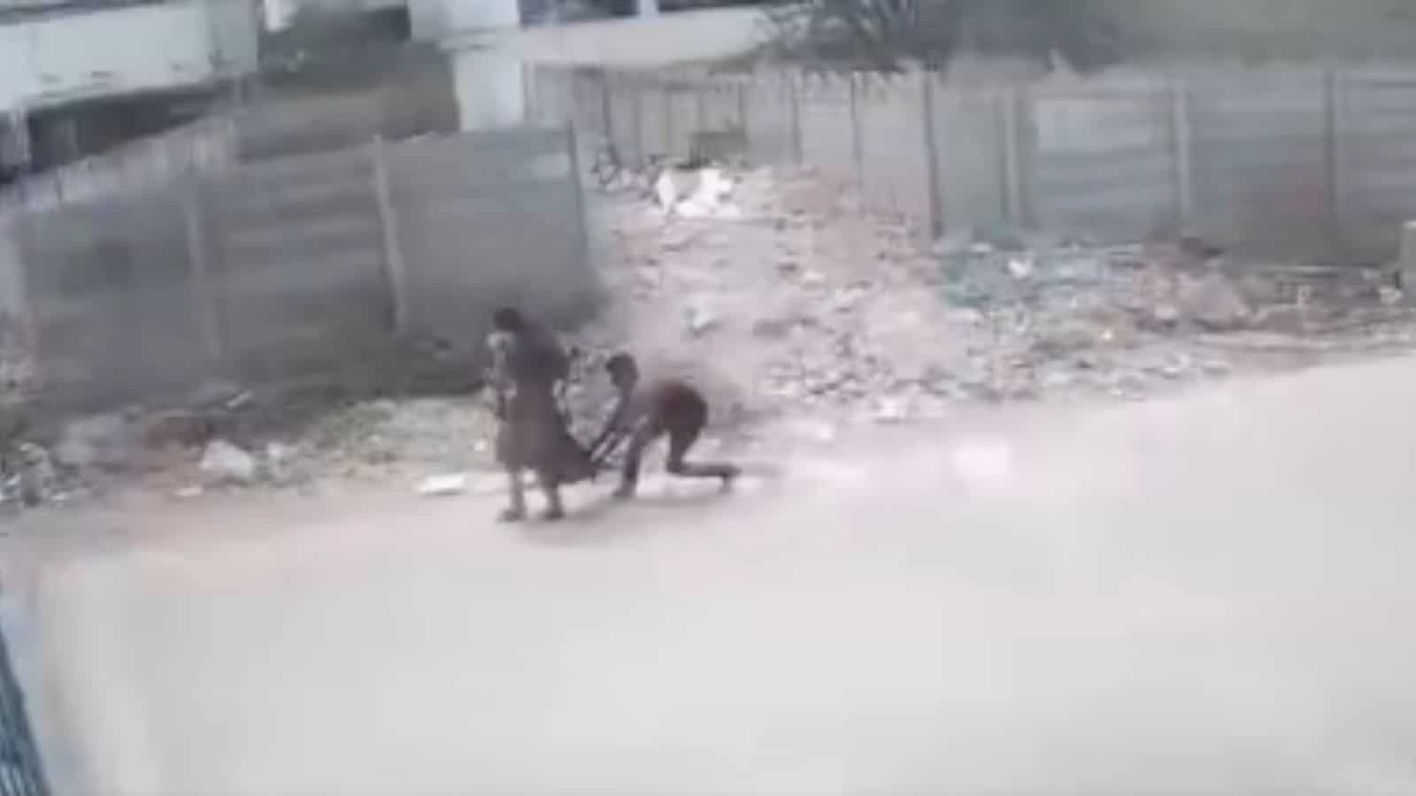 People walking along a dusty, unpaved road with scattered debris and fencing in the background.