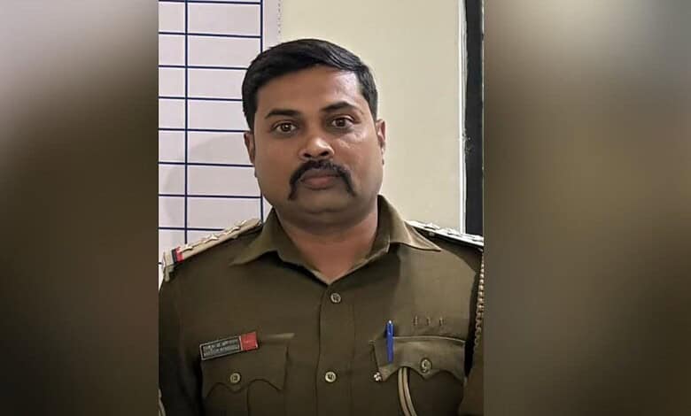 Police officer in uniform with badges and pens, standing indoors against a tiled wall.