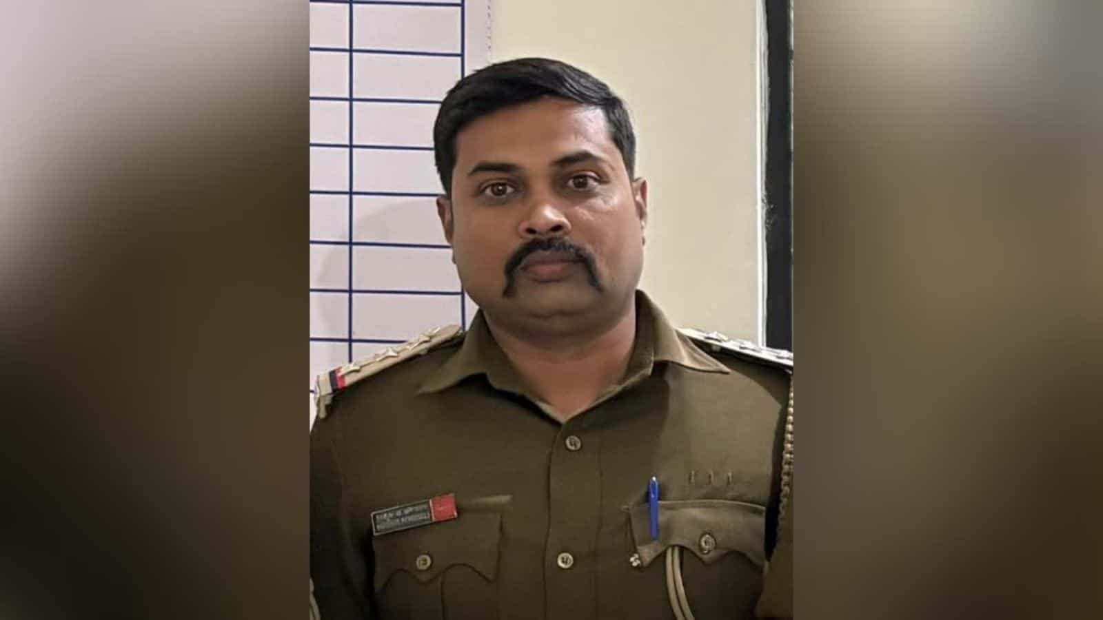 Police officer in uniform with badges and pens, standing indoors against a tiled wall.