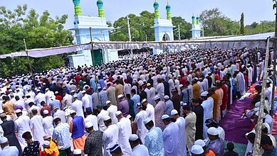 A woman leading prayer in a large outdoor congregation at a mosque with green minarets.