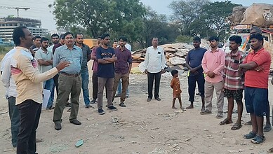A detailed group of people gathered outdoors, listening to a speaker in a dusty area with scattered debri.
