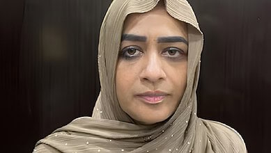 A woman wearing a beige hijab with a serious expression, indoors against a dark background.