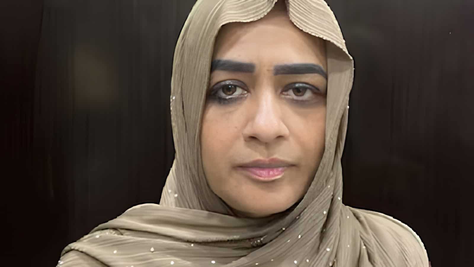 A woman wearing a beige hijab with a serious expression, indoors against a dark background.