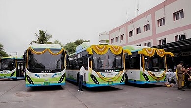 Modern electric buses decorated with yellow garlands parked outside a pink building in India.