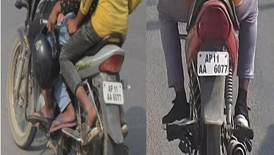 A man riding a motorcycle with a child sitting in front, both wearing helmets, on a busy street in India.