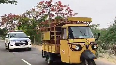 Yellow auto rickshaw on a rural road with a white car in the background, greenery and pink flowering tree.