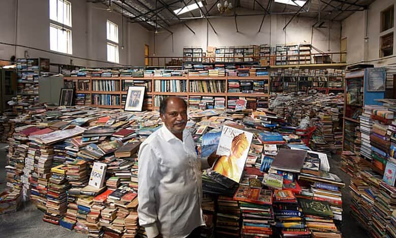 A busy warehouse filled with stacks of books and a man holding a book in the foreground.