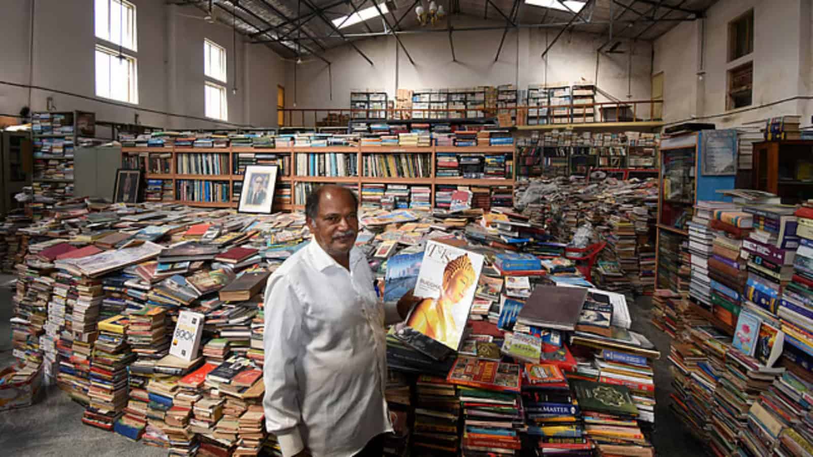 A busy warehouse filled with stacks of books and a man holding a book in the foreground.