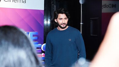 A young man with dark hair and a beard, wearing a blue sweater, standing at a media event or interview.