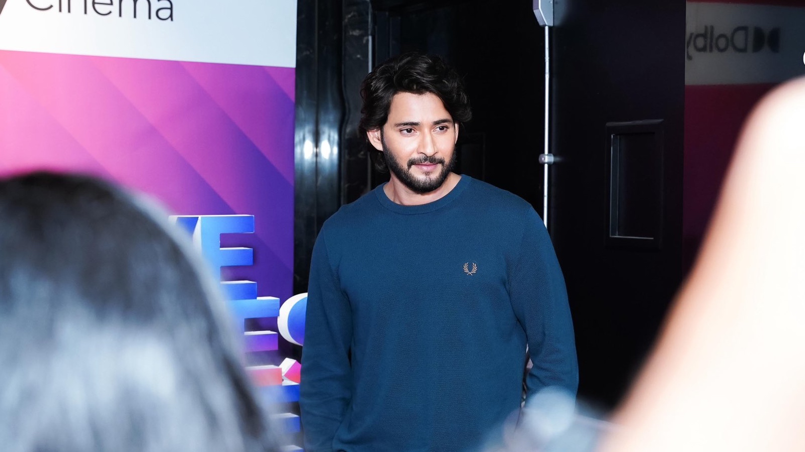 A young man with dark hair and a beard, wearing a blue sweater, standing at a media event or interview.