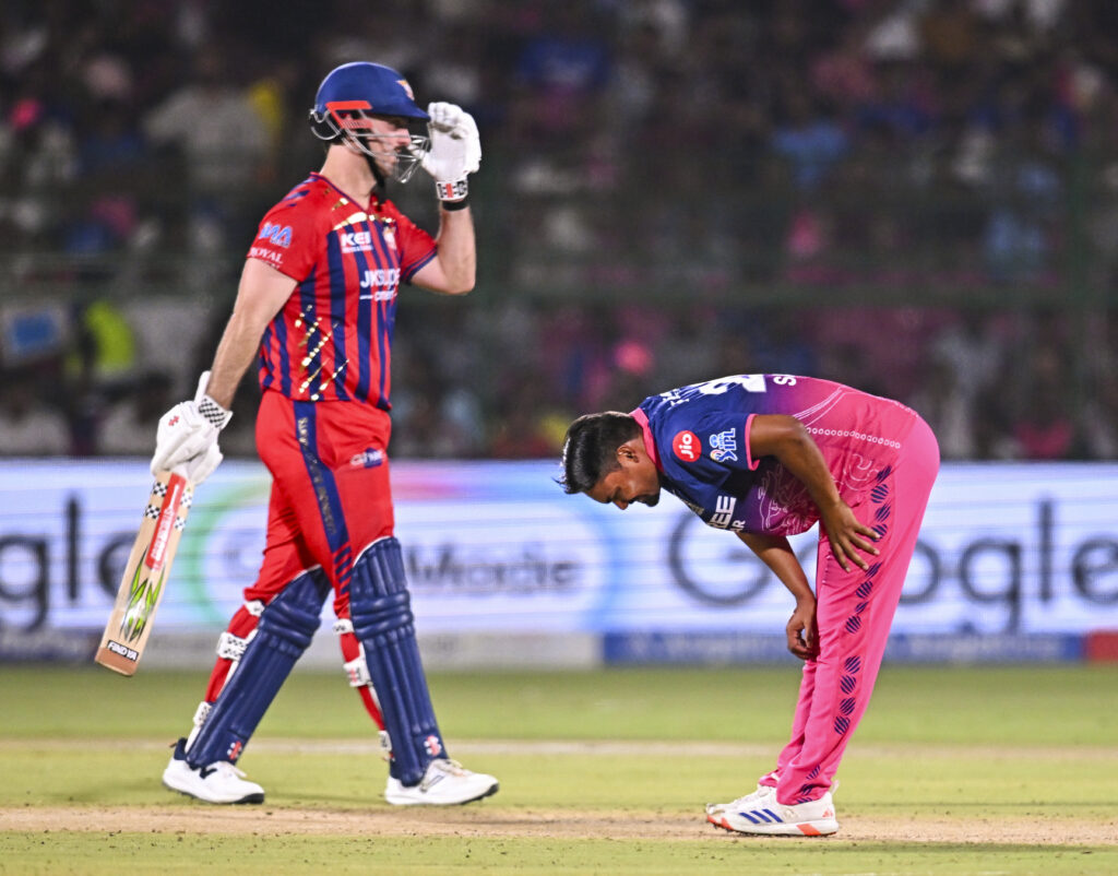 A cricket player in red and blue uniform with helmet and bat, and a player in pink uniform bowing on the.