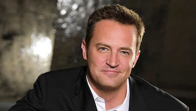 A man in a black suit smiling outdoors with sunlight filtering through trees.