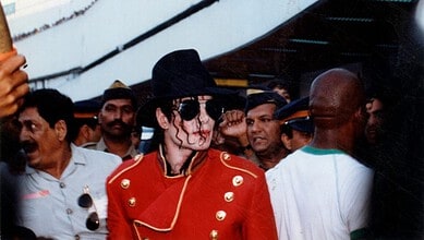 A woman dressed as Michael Jackson in a red jacket and black hat surrounded by a crowd at an event.