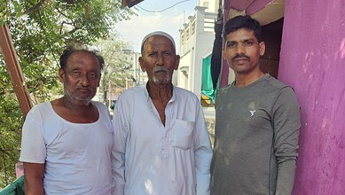 A man in a white kurta standing outdoors with two other men, one in a white t-shirt and the other in a gr.
