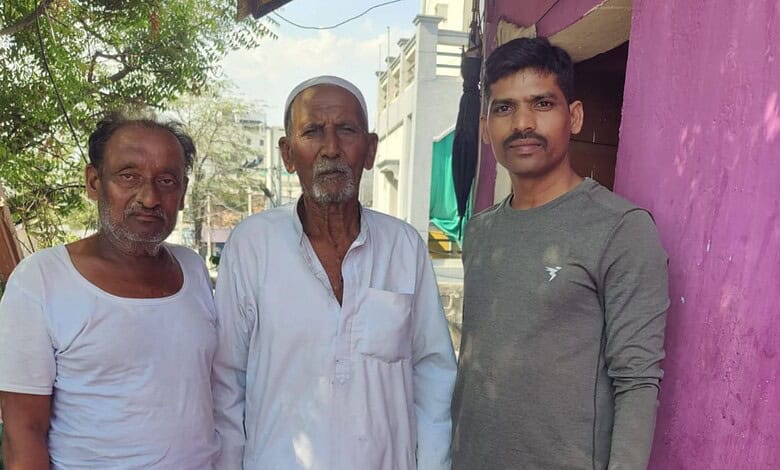 A man in a white kurta standing outdoors with two other men, one in a white t-shirt and the other in a gr.
