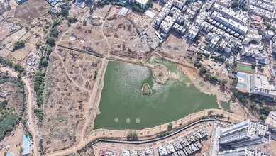 Aerial view of a park with a pond, surrounded by urban buildings and open land, in a cityscape.