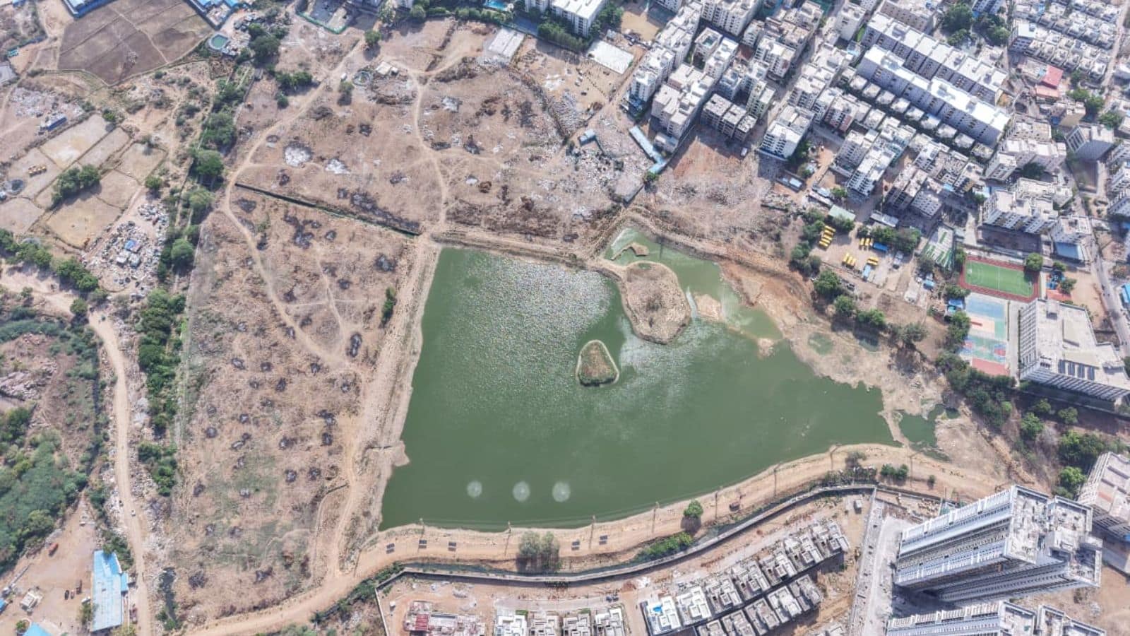 Aerial view of a park with a pond, surrounded by urban buildings and open land, in a cityscape.