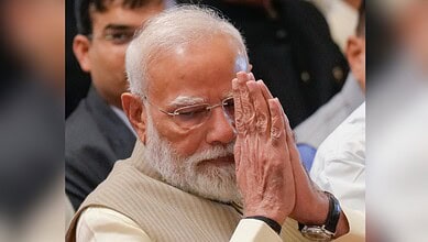 PM Narendra Modi praying with hands pressed together at a public event, surrounded by attentive attendees.