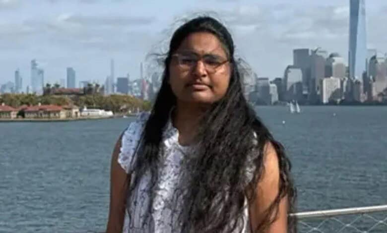 A woman with long black hair and glasses standing by the river with New York City skyline in the backgrou.