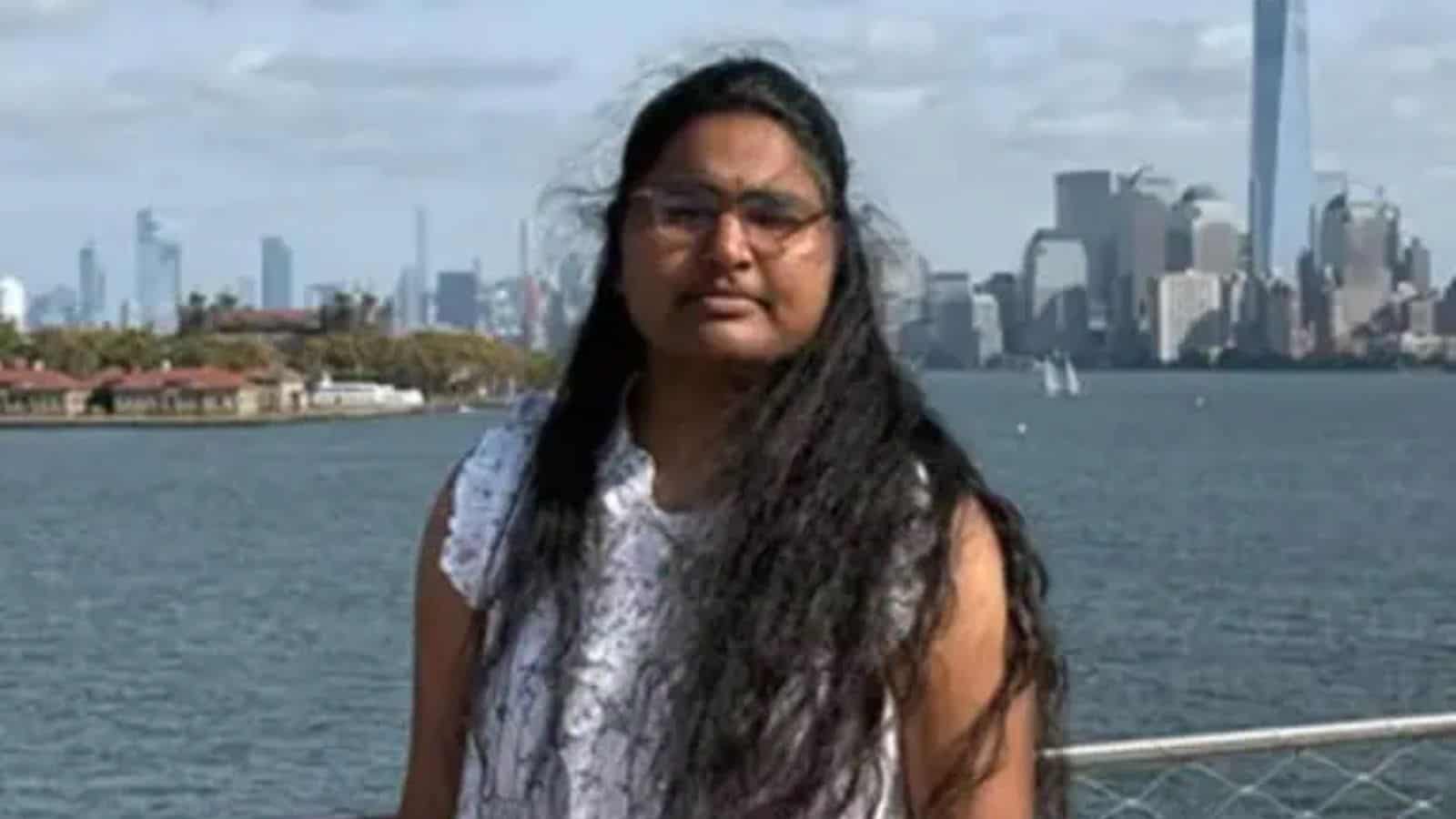 A woman with long black hair and glasses standing by the river with New York City skyline in the backgrou.