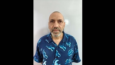Man with a beard and shaved head, wearing a blue shirt with cloud patterns, standing against a plain back.