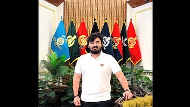 A man with a beard and white shirt standing in front of military flags in a formal setting.