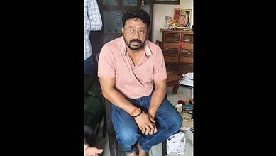 A man with glasses and a beard sitting on a chair in a cluttered room, wearing a pink shirt and blue jean.