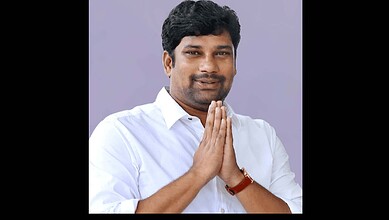 A person with dark hair and a white shirt holding hands in a traditional greeting gesture, smiling softly.