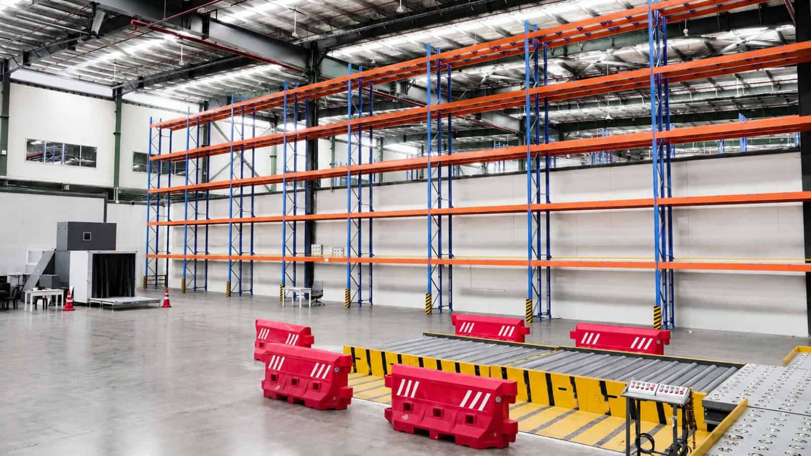 Aisle in a large warehouse with empty blue and orange shelving units and red safety barriers on the floor.