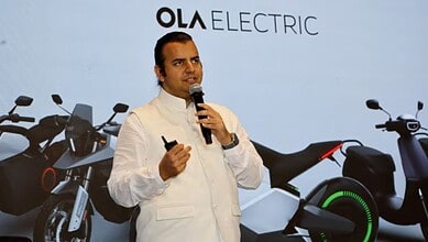 Man speaking at electric motorcycle launch event with Ola Electric branding and electric bikes in the bac.