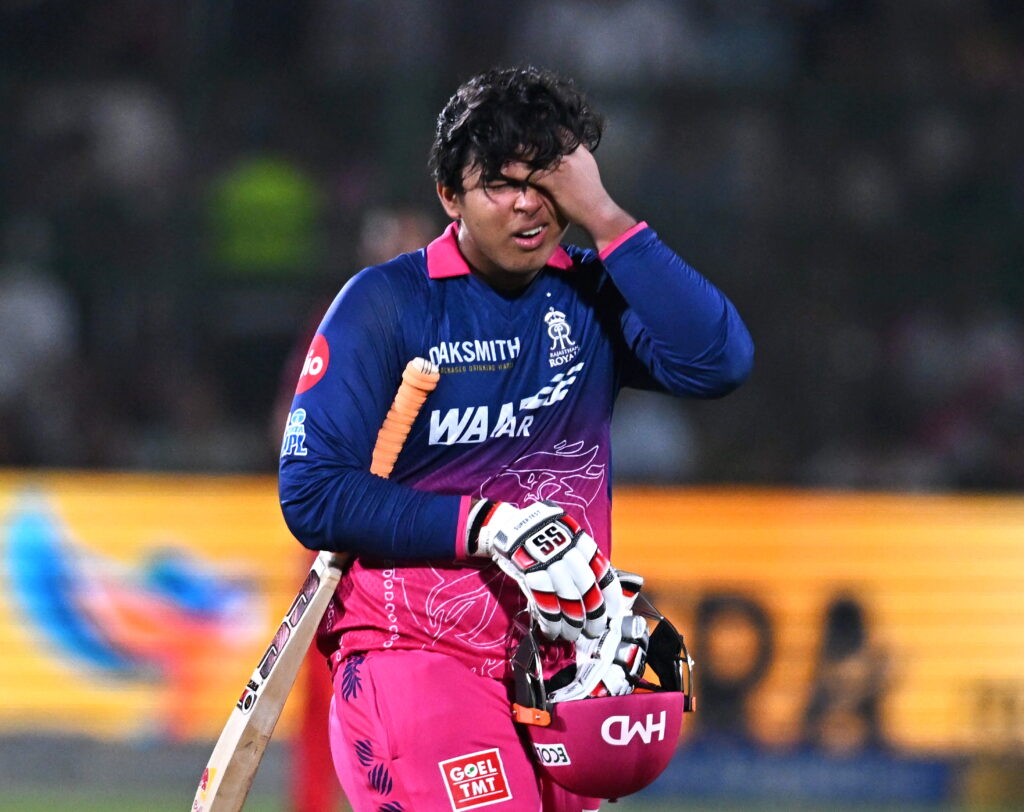 An athlete in pink and blue cricket uniform holding a bat, wiping sweat from his face during a match.
