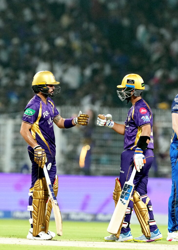 Two cricket players in purple and gold uniforms exchanging a fist bump on the field during a match.