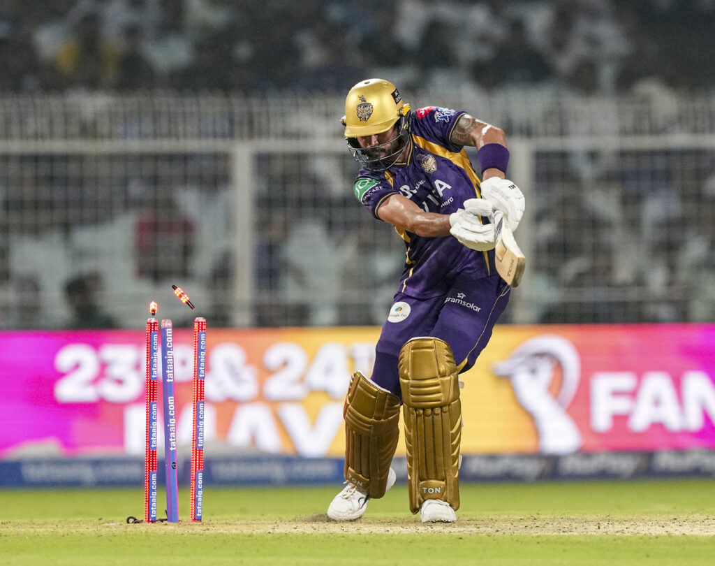 A1. ALT tag: Cricket wicketkeeper in purple and gold uniform preparing to catch the ball during match.