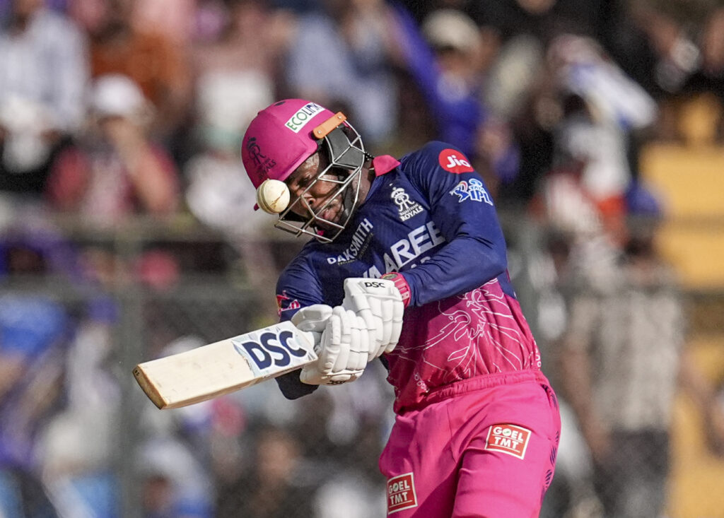 A woman cricketer batting during a match, wearing pink and blue uniform with helmet and gloves, in front.