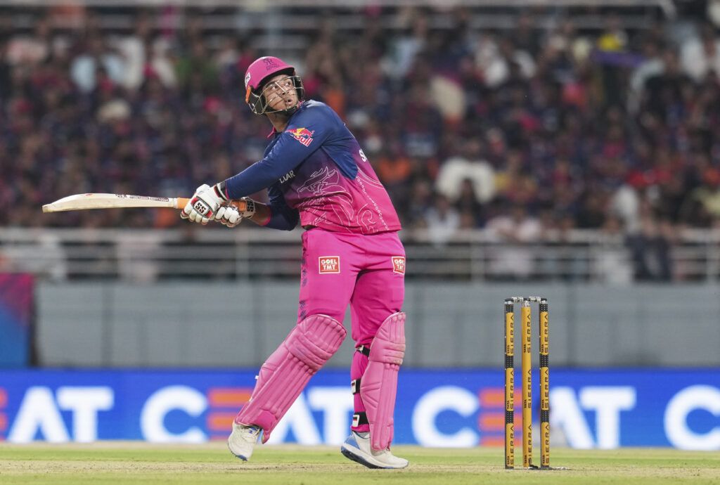 A woman cricket player in pink and blue uniform batting during a match on a cricket field.