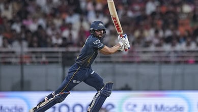 A young cricket player in action during a match, wearing a navy blue uniform and helmet, with a crowd wat.