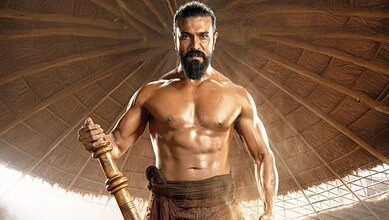 A muscular man with a beard, shirtless, holding a wooden staff in a traditional wooden structure, showcas.