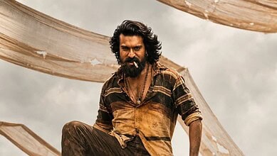 A man with long curly hair and a beard, dressed in rugged clothing, standing outdoors with fabric drapes.