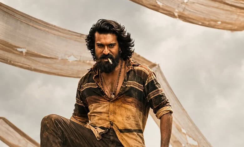A man with long curly hair and a beard, dressed in rugged clothing, standing outdoors with fabric drapes.