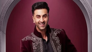 A smiling man dressed in a maroon embroidered sherwani, showcasing traditional South Asian fashion, stand.