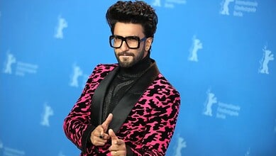 A man with dark hair and beard wearing black glasses and a pink and black patterned blazer at a media eve.