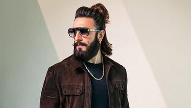 Man with long hair, beard, and sunglasses wearing a dark jacket and gold chain.