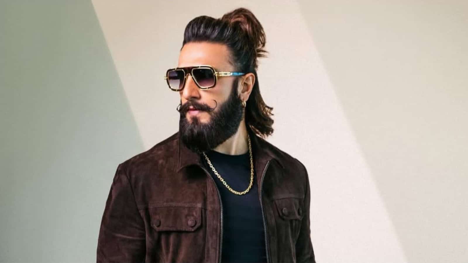Man with long hair, beard, and sunglasses wearing a dark jacket and gold chain.