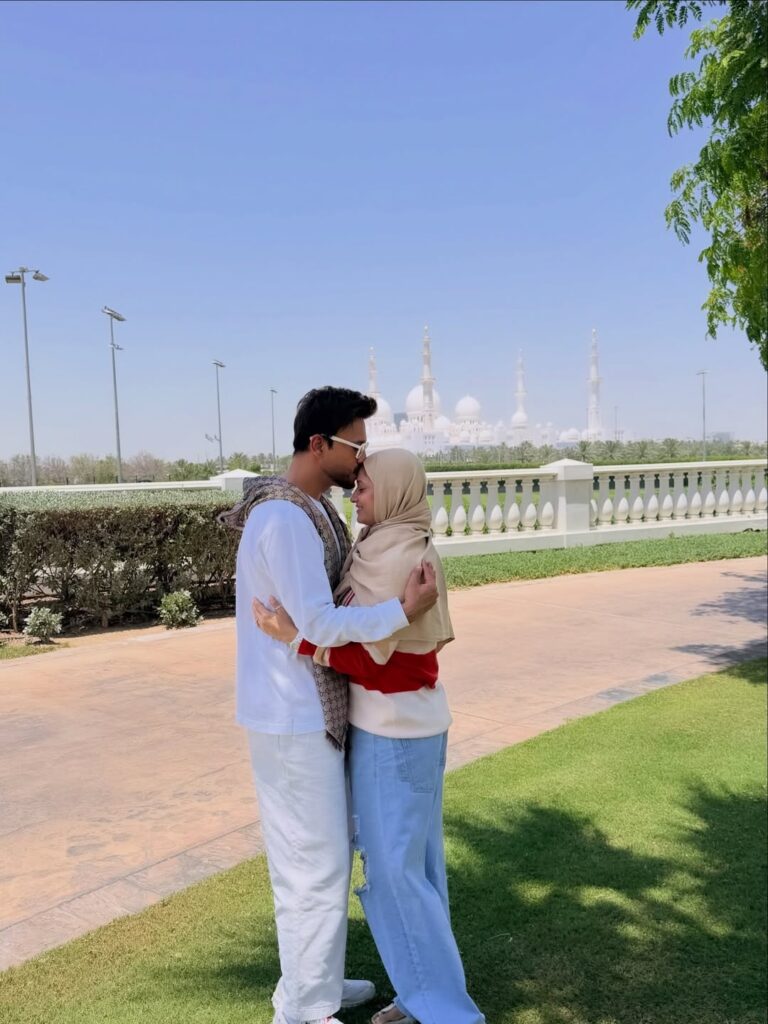A young couple sharing a tender moment outdoors with a mosque in the background, under a bright blue sky.