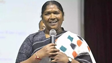Woman speaking into microphone at a press event, wearing a saree with a black and white pattern.